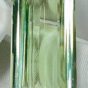 May include: A rectangular, faceted gemstone with a light green hue. The gem has a clear, transparent appearance with internal reflections. The cut features multiple facets, creating a play of light and shadow. The gemstone is set against a white background.