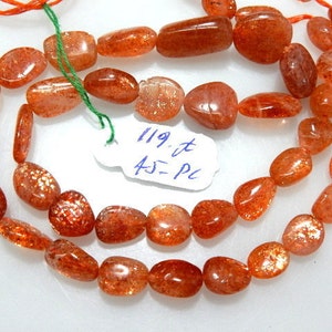 May include: Strands of polished, orange-red sunstone beads. The beads are oval and irregular in shape, with a sparkling, glittery effect. A small tag with handwritten text is attached to the strand.