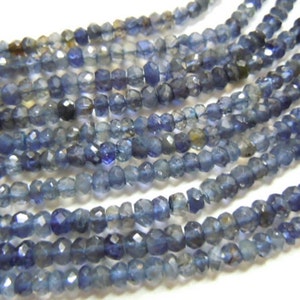 Puede incluir: Primer plano de varios hilos de cuentas facetadas de piedras preciosas azul-violeta. Las cuentas tienen una superficie pulida que refleja la luz. El color varía de índigo oscuro a tonos de azul más claros. Las cuentas están ensartadas, adecuadas para joyería.