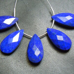 Puede incluir: Cinco piedras preciosas de lapislázuli facetadas en forma de lágrima ensartadas en un alambre de plata. Las piedras azul oscuro tienen una superficie pulida y reflectante, dispuestas en fila.