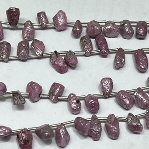 Peut inclure: Plusieurs brins de pierres précieuses de rubis brutes, de formes et de tailles variées. Les pierres sont d'un rouge-rose profond et sont enfilées sur un fil argenté. La texture rugueuse des pierres est visible, soulignant leur état naturel.
