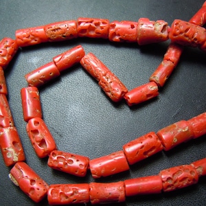 Puede incluir: Un collar de cuentas de coral rojo vibrante. Las cuentas cilíndricas presentan patrones tallados. El coral tiene un aspecto natural y texturizado, y las cuentas están ensartadas, creando un collar o pulsera.