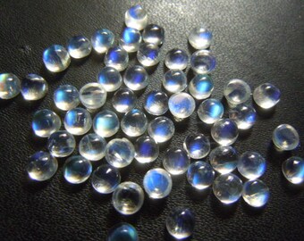 Puede incluir: Una colección de cabujones redondos de piedra lunar translúcida con un brillo iridiscente azul y blanco. Las piedras preciosas están dispersas sobre una superficie oscura, mostrando su forma pulida y abovedada. Ideales para la creación de joyas.