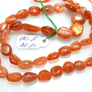 May include: Strands of polished, orange-red sunstone beads. The beads are oval and irregular in shape, with a sparkling, translucent appearance. A small tag with the text "121-dt A6-pc" is attached.