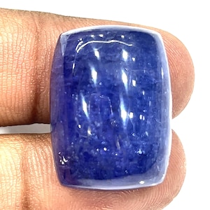 May include: A polished, rectangular gemstone with rounded edges. The gem is a deep, saturated blue color with a slight translucence. The surface reflects light, creating highlights. The gem is held by a person's fingers.