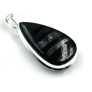 Peut inclure: Pendentif en forme de goutte d'eau avec une pierre noire rayée sertie dans un cadre en métal argenté. Le pendentif a une boucle en haut pour une chaîne. La pierre a une surface polie et réfléchissante.