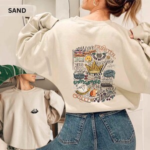 May include: A sand-coloured sweatshirt with a graphic design on the back, including a crown, a guinea pig, and various phrases. The sweatshirt has long sleeves and a crew neck. The word "SAND" is printed on the top left.