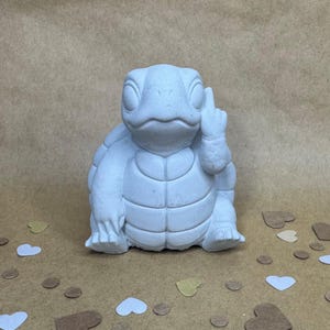 May include: A light gray turtle figurine with a hand gesture. The turtle is sitting upright, with its head and limbs visible. The figurine is made of a solid material and has a textured surface. Decorative hearts and circles are scattered around the base.