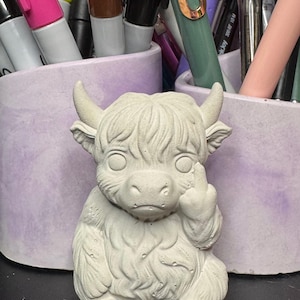 May include: A light gray concrete Highland cow figurine, seated with one hand raised in a rude gesture. The cow has small horns and a shaggy coat. The figurine is in front of a lavender pen holder filled with colorful pens and markers.