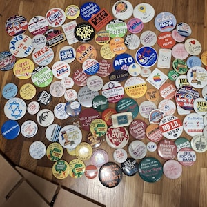 May include: A collection of colorful vintage pinback buttons with various designs and text, including phrases like "Quick Fixin' Ideas," "I Care," and "Free." The buttons are scattered on a wooden surface.