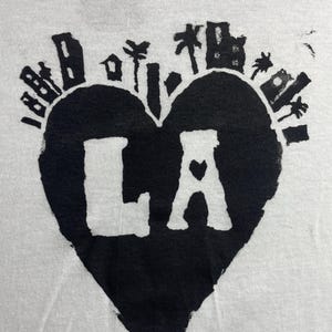 May include: White t-shirt with a black heart-shaped graphic. The graphic features the letters "LA" with a heart in the "A", and a cityscape and palm trees around the heart. The shirt has a slightly worn appearance.
