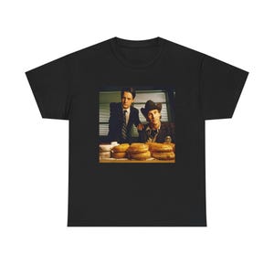Puede incluir: Camiseta negra con un gráfico de dos hombres y donuts. Uno de los hombres lleva un traje, el otro un sombrero de vaquero. La imagen es una referencia a la serie de televisión Twin Peaks. Los donuts son un elemento clave.