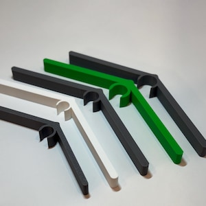 Może przedstawiać: Asortyment plastikowych klipsów do toreb w różnych kolorach, w tym białym, ciemnoszarym i zielonym. Klipsy mają zakrzywioną konstrukcję do zamykania toreb i są ułożone na jasnoszarej powierzchni.