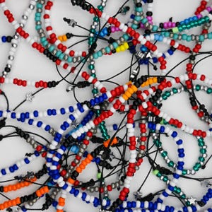 May include: A collection of colorful beaded bracelets in various colors including red, white, blue, orange, and black. Some bracelets feature small star-shaped charms. The bracelets are strung on black cords.