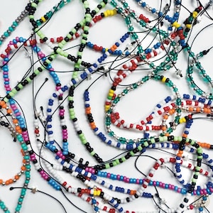 May include: A collection of colorful beaded bracelets in various colors including red, green, blue, and silver. The bracelets are strung on black cords and feature small star charms. The beads are small and round.