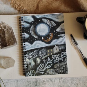 May include: A spiral-bound notebook featuring a dark, nocturnal illustration of a full moon, a flying owl, a leaping fox, and a small house surrounded by bare trees. The cover art uses shades of black, white, and blue to depict a night scene.