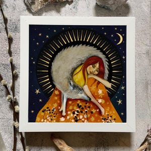 May include: A square framed print featuring a person with red hair resting against a large grey wolf. The background is a dark blue night sky with a golden crescent moon and stars, framed by a circular golden sunburst design over an orange field.