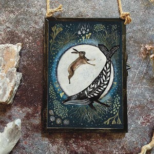 Op de afbeelding: Een ingelijste kunstdruk met een bruine haas die voor een walvis springt met een zwart-wit patroonstaart. Het kunstwerk is geplaatst tegen een donkerblauwe achtergrond met botanische elementen en een witte cirkel. De lijst is donkerbruin.