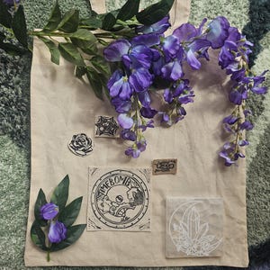 May include: A beige canvas tote bag decorated with a sprig of purple wisteria flowers, a small black and white rose patch, a square patch with the text TIMEBOMB, a small rectangular patch, and a square coaster featuring a white crescent moon and crystal design.