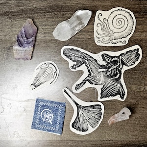 Könnte beinhalten: Eine Ansammlung natürlicher und künstlerischer Objekte, darunter ein lila-weißer Kristall, ein klarer Quarzkristall und mehrere Schwarz-Weiß-Linolschnitte eines Nautilus, eines Fledermaus-Skeletts, eines Trilobiten und eines Ginkgoblattes. Ein kleines Jeans-Patch mit weißen Nähten und ein geschnitzter Vogelschädel sind ebenfalls vorhanden.