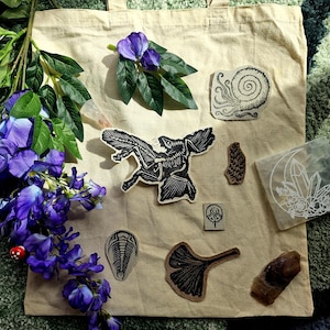 May include: A beige canvas tote bag decorated with several black and white patches featuring a bat skeleton, a spiral shell, a trilobite, a ginkgo leaf, and a crystal design. A sprig of purple wisteria flowers is placed on the left side of the bag.