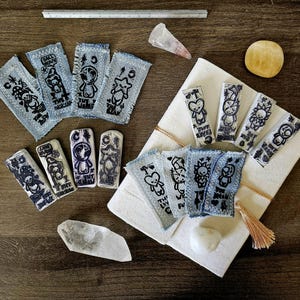 Pode incluir: Uma variedade de peças de arte feitas à mão, incluindo patches de ganga com ilustrações a preto e branco, selos de cerâmica e cristais. Os patches apresentam vários personagens de desenhos animados e texto. Os itens estão dispostos em uma superfície de madeira.