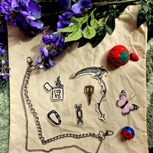 May include: A collection of items including a silver metal chain with lobster clasps, several small fabric patches featuring black ink illustrations of a scythe, a lighter with a tooth, scissors, a carabiner, and a chain link, a strawberry pincushion, and a butterfly.
