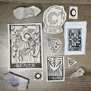 Puede incluir: Una colección de artículos espirituales y ocultos, que incluyen parches de cartas del tarot, piedras de cristal y una planchette. Los parches presentan diseños de linograbado de una bola de cristal, la carta de la Muerte, la carta de la Estrella y Tres de Espadas.