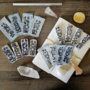 May include: A collection of handmade art pieces, including denim patches with black and white illustrations, ceramic stamps, and crystals. The patches feature cartoon characters and text. The items are arranged on a wooden surface.