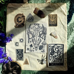 May include: A beige canvas tote bag featuring a central rectangular patch with a deer skull, moth, and mushroom illustration. Surrounding patches include a crystal ball, a planchette, the 3 of Swords, The Star, and Death tarot cards.