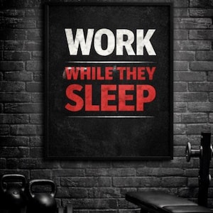 May include: A framed black poster with the words "WORK WHILE THEY SLEEP" in white and red. The poster is hanging on a dark brick wall, with gym equipment visible in the foreground, including kettlebells and a weight bench.