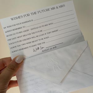 Può includere: Una carta bianca con il testo "WISHES FOR THE FUTURE MR & MRS" e domande da completare. La carta è all'interno di una busta bianca con iniziali in rilievo e la data 22 agosto 2025.
