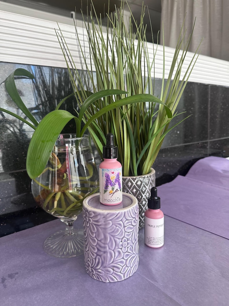 May include: Two pink dropper bottles with black lids and white labels, one with the text "moochte" and a lavender sprig design, the other with "TRIPLE PEPPER". They are on a lavender floral ceramic holder, with a glass vase and potted plants in the background.