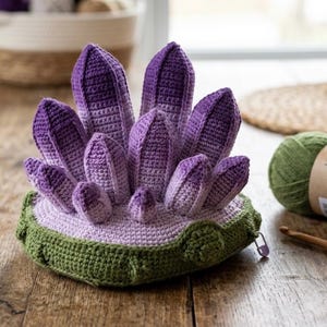 Peut inclure: Une pièce amigurumi crochetée à la main représentant un groupe de formations cristallines pointues en dégradé de violet à lavande, émergeant d'une base circulaire violet clair avec un bord extérieur texturé vert olive foncé et une petite fermeture éclair.