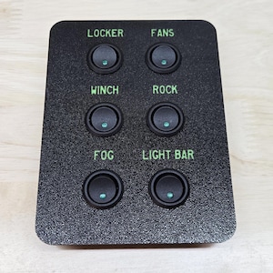 May include: A black control panel with six illuminated rocker switches. The switches are labeled in green with the following text: LOCKER, FANS, WINCH, ROCK, FOG, and LIGHT BAR. The panel is rectangular and sits on a light wood surface.