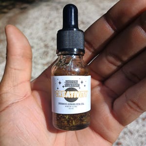 May include: A small glass bottle of "Creativity" Voodoo Condition Oil, held in a hand. The bottle has a black dropper top and a white label with text and star designs. The oil inside contains herbs. Made in the USA.