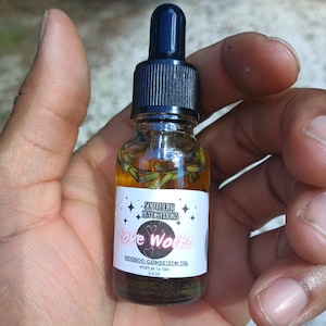 May include: A small glass bottle of Hoodoo Condition Oil, held in a hand. The bottle has a black dropper top and a white label with the text "Southern Invocations Love Works". The oil inside contains green herbs.
