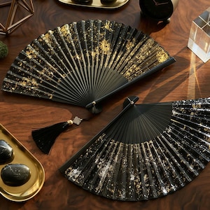 May include: Two black hand fans with gold leaf accents are displayed on a wooden surface. The fans are open, showcasing their intricate design. A black tassel hangs from one fan. Decorative items like a clock and stones are also present.