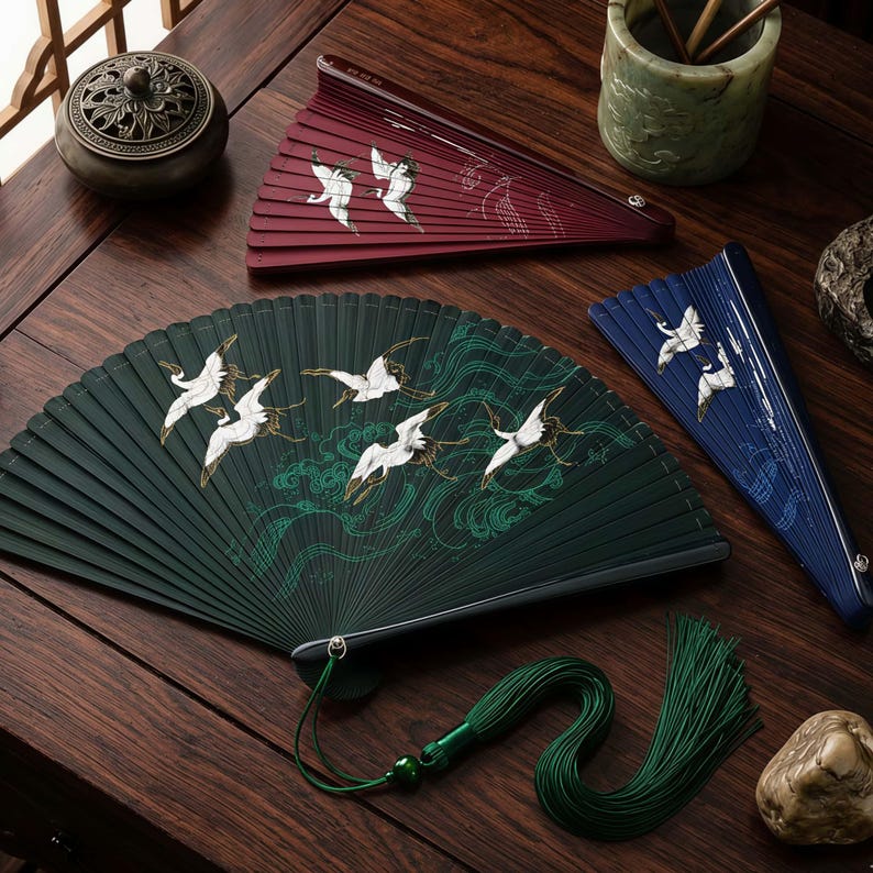 May include: A selection of decorative hand fans in green, burgundy, and blue. Each fan displays a design of white cranes in flight above stylized waves. A green tassel hangs from one fan, adding a decorative element.
