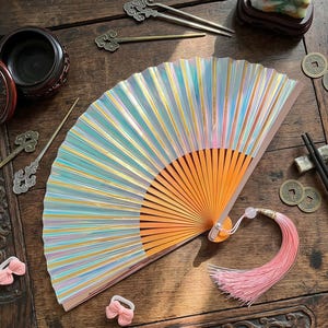 May include: Iridescent hand fan with a peach tassel and a wooden handle. The fan has a rainbow effect with gold accents. The fan is open, displaying its full shape. Other Asian-inspired decorative items are on the wooden surface.