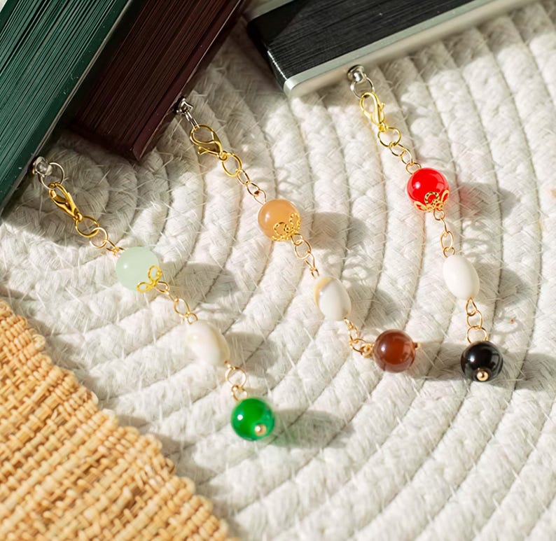 May include: Three gold-toned metal chains attached to rectangular objects, each featuring a series of colorful beads. The beads include shades of light green, white, orange, red, brown, and black, with some beads accented by small gold-toned metal caps.