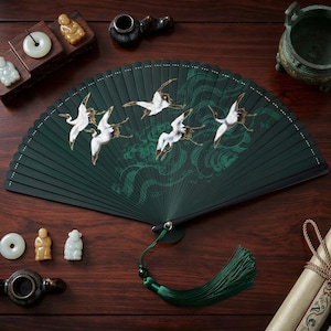 May include: A dark green folding fan with a crane design. The fan features white cranes in flight over green wave patterns. A green tassel hangs from the base. Additional Asian-inspired decor surrounds the fan on a wooden surface.