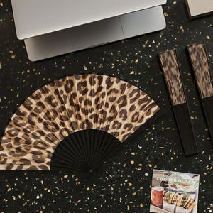 May include: An open hand fan with a leopard print design and a black handle. Two closed fans with a similar design are next to it. A laptop and a magazine are also on a black surface.