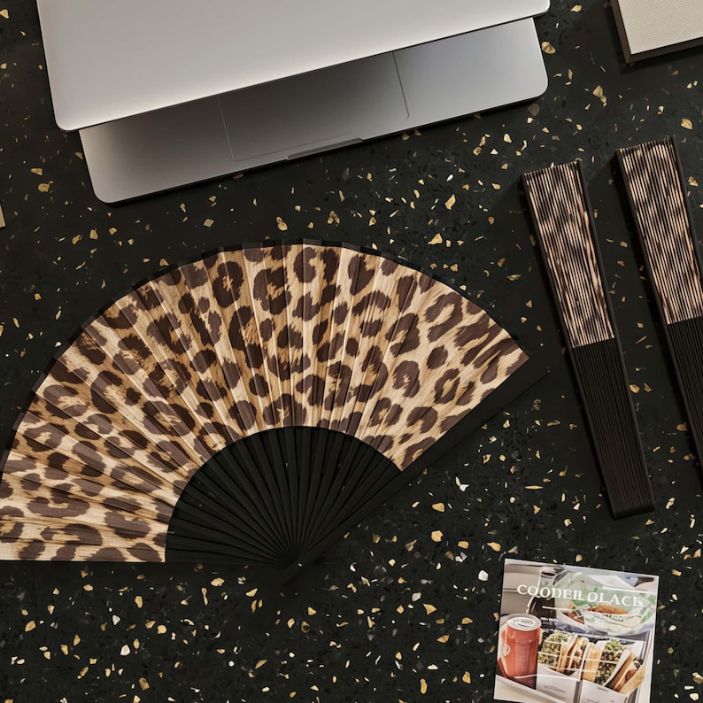 May include: An open hand fan with a leopard print design and a black handle. Two closed fans with a similar design are next to it. A laptop and a magazine are also on a black surface.