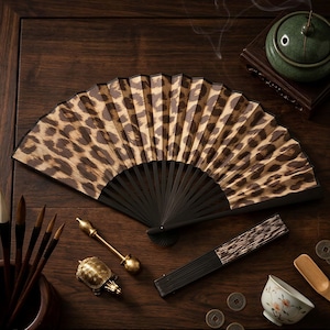 May include: An open leopard print folding fan with black ribs rests on a dark wooden surface. Other items include calligraphy brushes, a small turtle figurine, a small teacup, and a green incense burner.