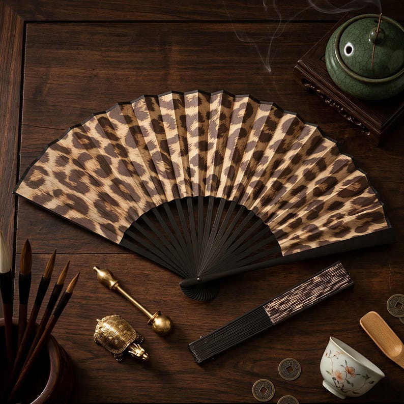 May include: An open leopard print folding fan with black ribs rests on a dark wooden surface. Other items include calligraphy brushes, a small turtle figurine, a small teacup, and a green incense burner.