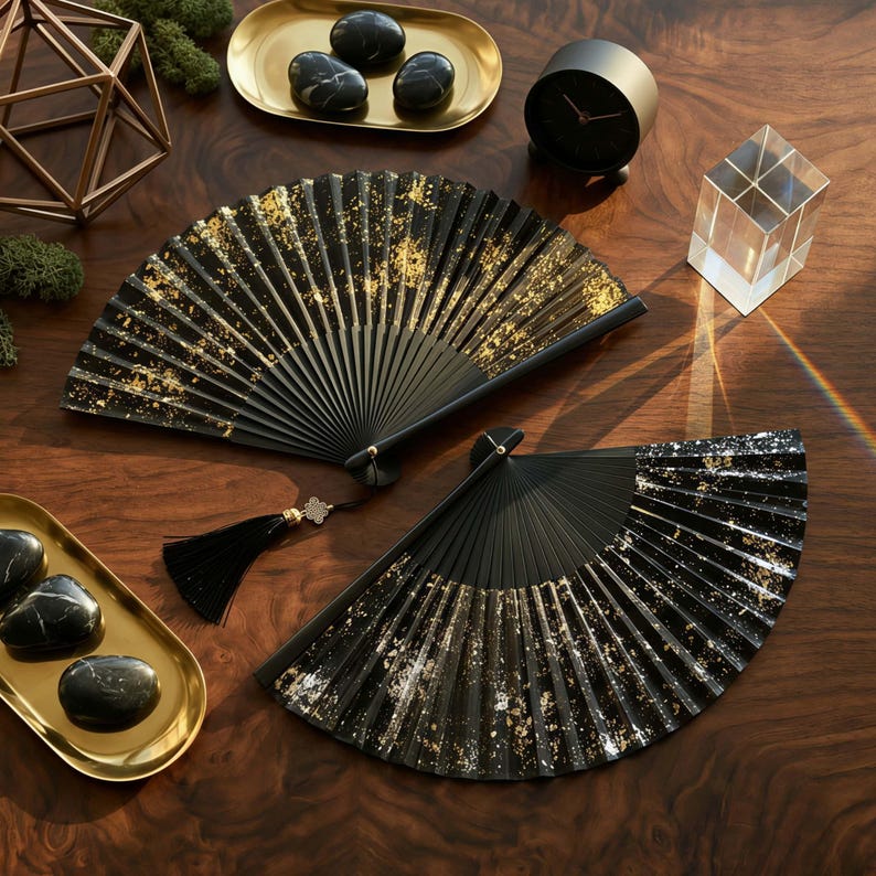 May include: Two black hand fans with gold leaf accents are displayed on a wooden surface. The fans are open, showcasing their intricate design. A black tassel hangs from one fan. Decorative items like a clock and stones are also present.