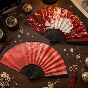 Può includere: Due ventagli pieghevoli decorativi con manici neri. Uno presenta un disegno di volpe rossa e bianca, l'altro un motivo floreale rosso. I ventagli sono su una superficie di legno scuro con accenti decorativi.