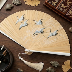 May include: A beige bamboo hand fan with a white handle and tassel. The fan features a painted design of white cranes in flight against a backdrop of stylized clouds. The fan is a traditional Asian art form.