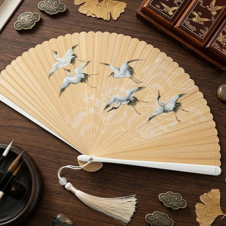 May include: A beige bamboo hand fan with a white handle and tassel. The fan features a painted design of white cranes in flight against a backdrop of stylized clouds. The fan is a traditional Asian art form.
